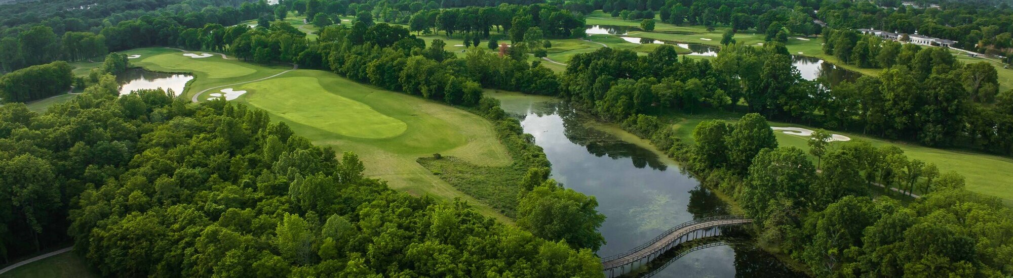 Aerial view of a lush green golf course with a pond and a wooden bridge surrounded by trees.
