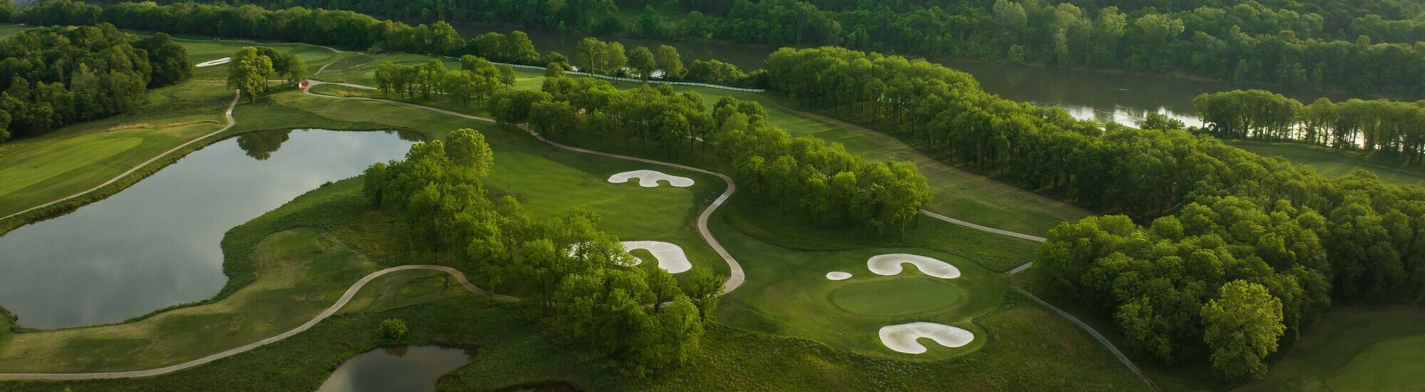 Aerial view of a lush golf course with sand traps, a pond, and tree-lined borders under a clear sky.
