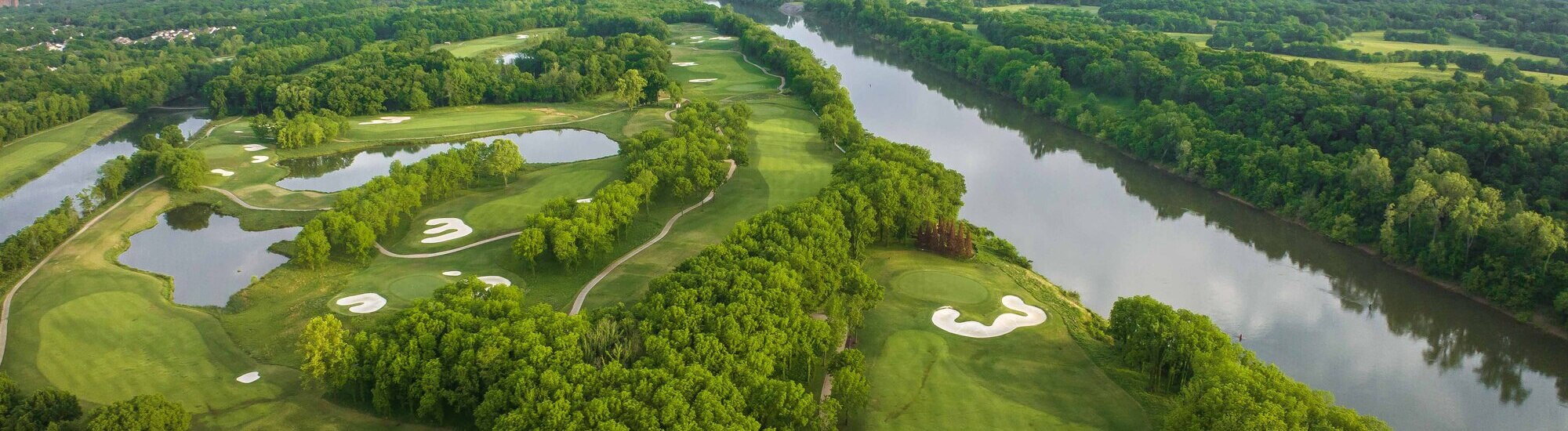 Aerial view of a lush green golf course with winding paths and a river, surrounded by dense trees.