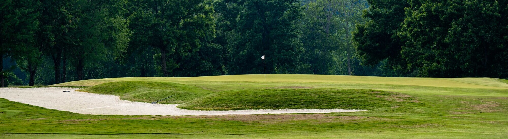 A golf course landscape featuring a sand trap, green grass, and a flag on the putting green in the background.