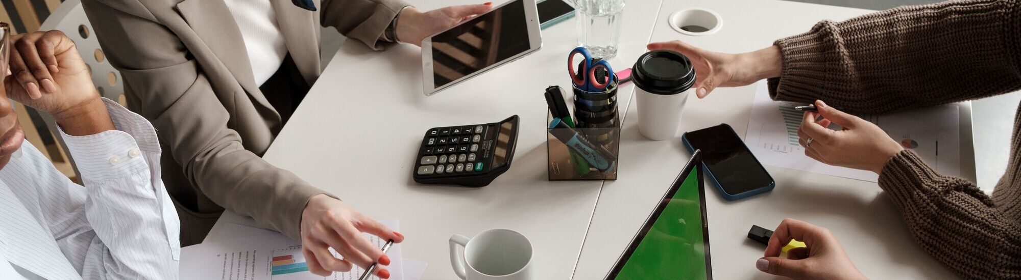 A meeting table with hands gesturing, a calculator, tablets, and stationery items, indicating a collaborative discussion.