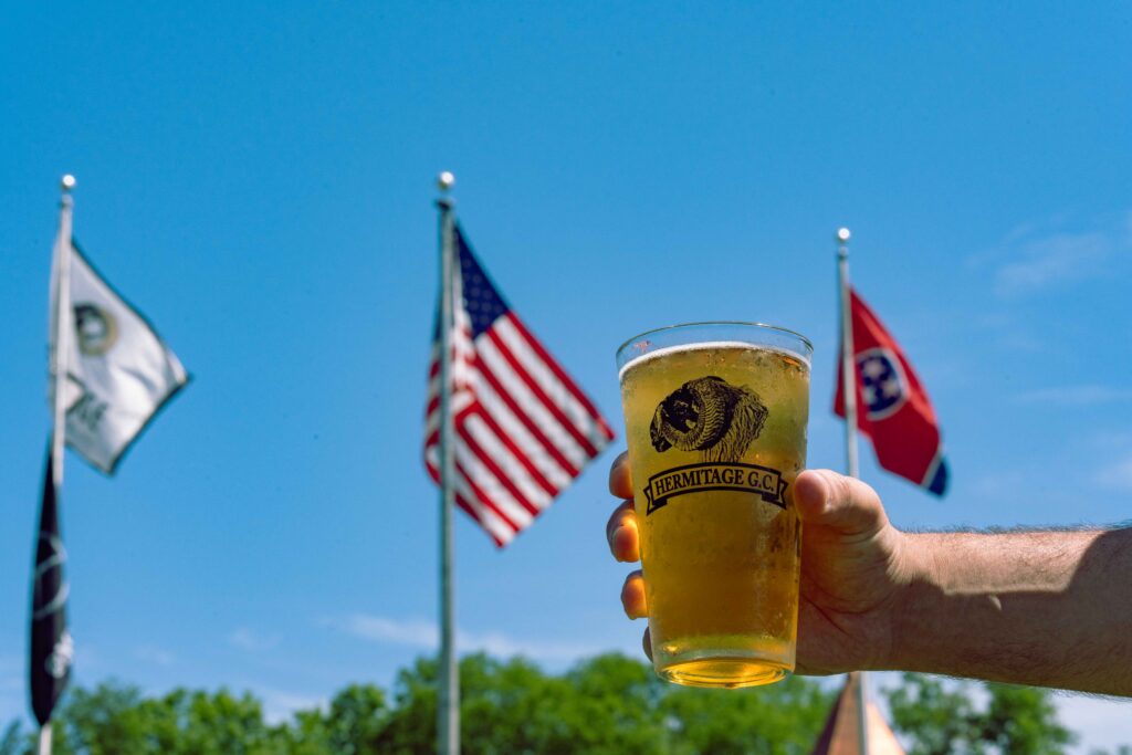 A hand holding a glass of beer with 'Hermitage G.C.' logo, flags in the background against a clear blue sky.