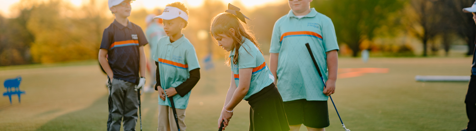 A group of children practicing golf on a sunny day, focusing on a young girl preparing to putt.