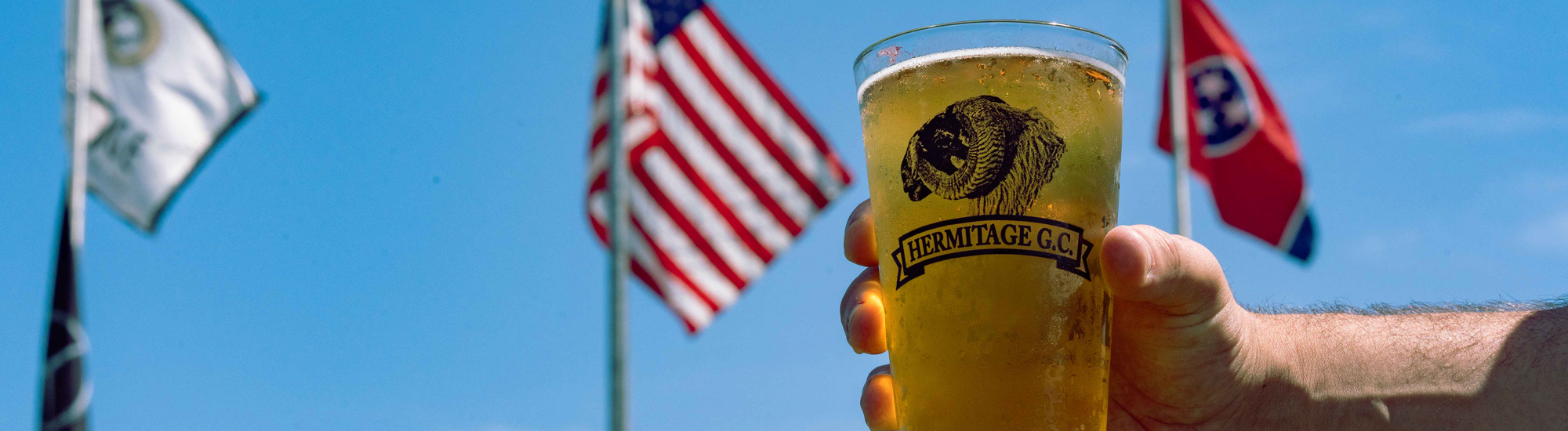 A hand holding a glass of beer with a logo, flags in the background against a clear blue sky.