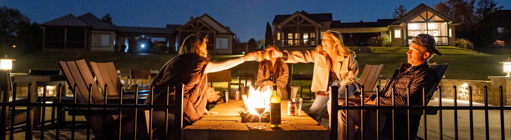 Group of friends enjoying a fire pit at night, sharing drinks and laughter in a cozy outdoor setting.