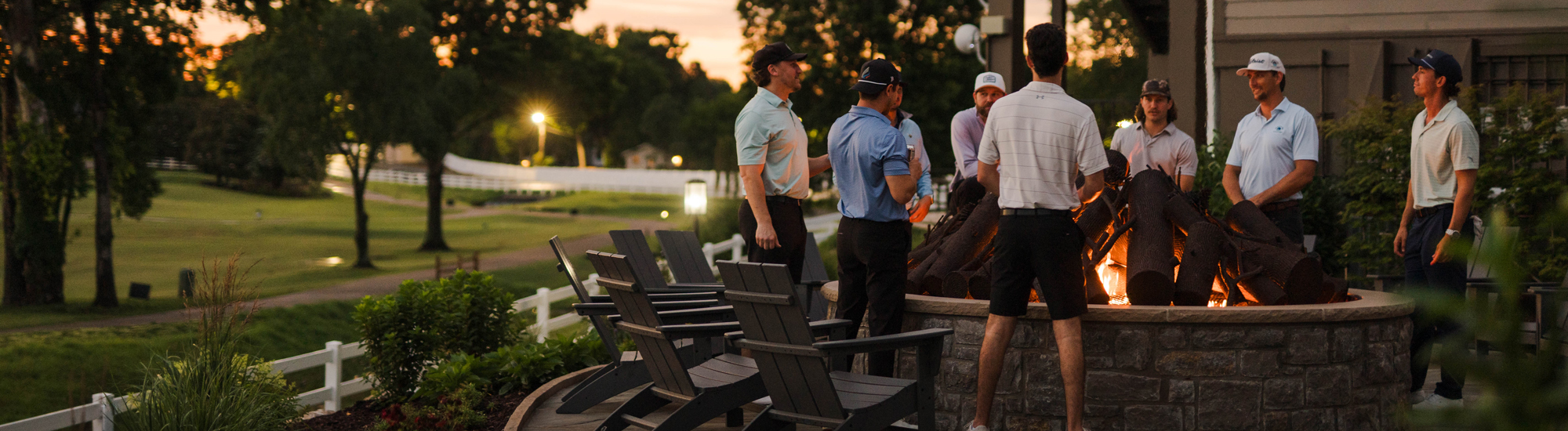 Group of people gathered around a fire pit at dusk, with a golf course in the background.