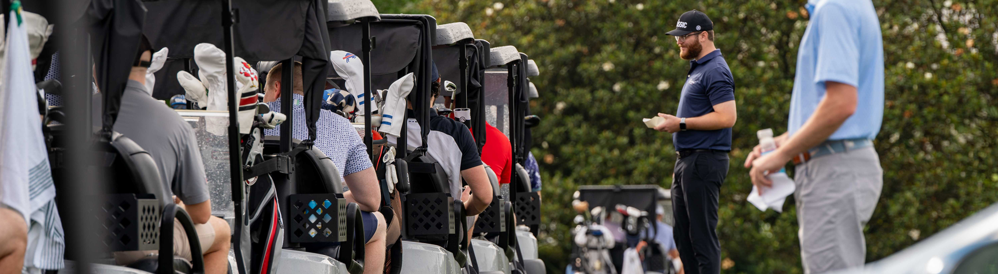 Golfers seated in carts with clubs, while a man in a blue shirt speaks to them on a tournament day.